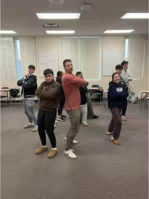 A group of people, likely high school students and a teacher, are posing in a classroom. The teacher and several students cross their arms in front of their chests, in a pose. The lighting is bright, and the room appears to be a classroom with windows. The overall impression is of teamwork and a shared goal.