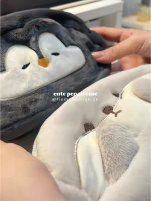 The image shows a close-up of two plush penguin pencil cases; one is dark gray with white penguin faces, and the other is white. A person's hand can be seen holding the cases, with the focus on the cute, fluffy details of the penguin designs.