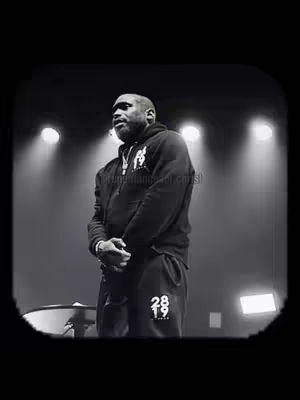 A black and white photo shows a man standing on a stage in front of bright stage lights. He wears a black hoodie and sweatpants displaying the number 2819. His hands are clasped in front of him, and he appears to be looking downwards. The background is dark and out of focus.
