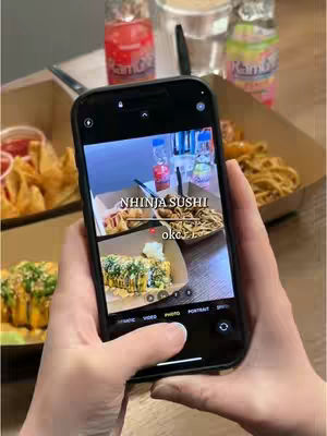 A person is holding a phone which is displaying a photo of food from a sushi restaurant. The phone is angled towards the viewer. The person's hands are visible, holding the phone. In the background are items associated with a meal: food, drinks, and a table setting.