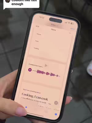 A close-up shot of a woman holding a smartphone. The phone's screen displays an app interface with text and a waveform visualization, suggesting an audio recording or editing application. The background is blurred, with details like a glimpse of a white shoe visible.