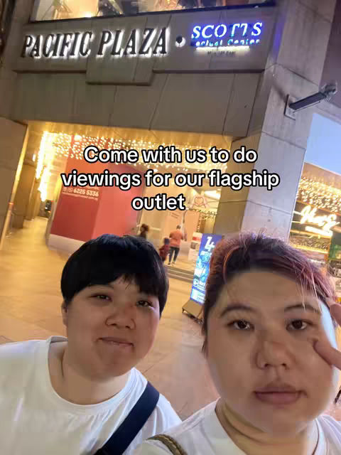 A selfie-style image featuring two women in front of a building. The building's signage includes 'PACIFIC PLAZA' and 'SCOTTS'. The women are smiling, and overlaid text at the bottom reads 'Come with us to do viewings for our flagship outlet'.