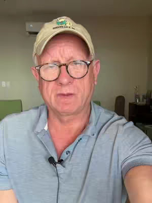 A man wearing glasses and a baseball cap is looking at the camera in front of a background with a light-colored wall and AC unit. He is wearing a light blue polo shirt. A microphone is clipped to his shirt.