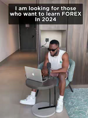 A man in a white tank top and sunglasses is seated in hotel room, with a laptop and microphone on a small table in front of him. A text overlay states 'I am looking for those who want to learn FOREX In 2024'. The background shows a hallway with a closed door and a carpeted floor.