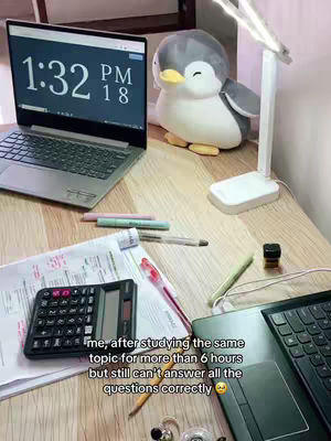 A top-down shot of a wooden desk with a laptop showing 1:32 PM on the screen, a penguin plushie, and a desk lamp. Various stationery like pens, a calculator, and printed papers are scattered around. Text overlays are present.