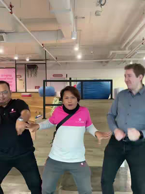 The cover image shows three people, presumably in an office or similar indoor space. The main focus is a person in the center wearing a pink and white shirt and grey pants, with two other people on either side. The central person is the focal point and has their arms out and is dancing with the other two mirroring their pose. The setting is nondescript with some open space, a drop ceiling and some blue bean bag type furniture in the background.