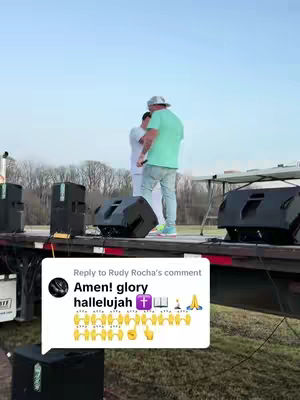 Two men stand on a stage setup on a flatbed truck, addressing an audience. Speakers and equipment surround them under a clear sky. A comment section overlay includes text and emojis. 