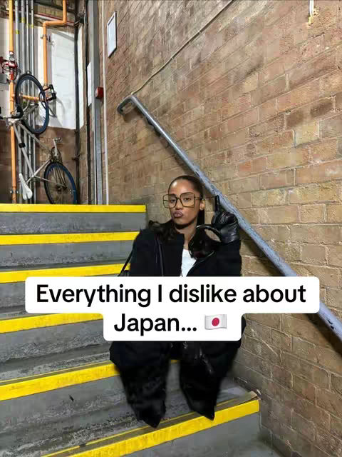 A person with glasses and a black outfit is looking directly at the camera and holding their right hand up with an object that has a Japanese flag on it. They are sitting on some stairs which is made of yellow and grey bricks and the background behind them is from some building structure.