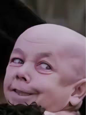 Close-up of a pale, unsettling face resembling a baby, with exaggerated features and a mischievous expression. The eyes are looking askance, and the mouth is slightly open in a smirk. The background is blurred but appears to be a dark, interior space.