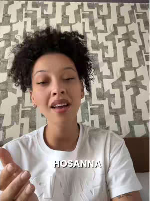 A woman with curly dark hair is centered, looking slightly upward. She is wearing a white t-shirt, and a text overlay of the word "HOSANNA" is in a white rectangular box beneath her chin. Behind her is a wall with a patterned design.