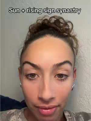 A selfie-style shot focused on a woman's face, looking at the camera with a neutral expression. Her hair is pulled back in a bun. The background is a simple, light-colored wall. Text overlay indicates the video's topic: "Sun + rising sign synastry".