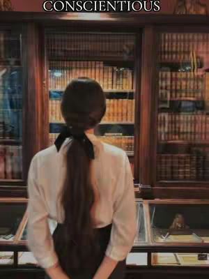 A woman from the back is standing in a library, dressed in a white shirt and black skirt. Her hair is long and pulled back with a black ribbon. The background is filled with bookshelves.