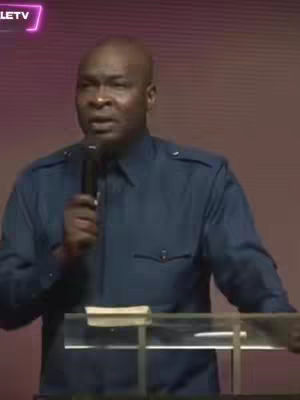 A man with a bald head and blue collared shirt is speaking into a microphone, standing behind a clear podium with a book on top. Behind him is a plain background with a subtle pattern, and a logo is visible in the top left.
