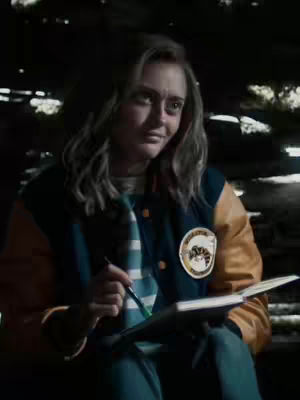 A medium shot features a person with long, wavy blonde hair, wearing a blue and yellow varsity jacket with a patch. The person is holding a pen and notebook, seemingly concentrating. The background appears dark and out of focus, possibly an exterior setting.