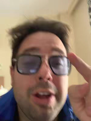 A close-up shot of a man wearing large-frame dark sunglasses, pointing a finger towards the side of his head. He has short, dark hair and a bit of stubble, and is wearing a blue collared shirt. The background is softly lit, with hints of a wall and curtains.