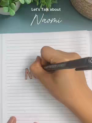 The cover image shows a hand writing on a lined notebook. The hand holds a black pen, and is in the process of coloring in the letter "N" of the word "Naomi". The background is a mix of surfaces, including a greenish surface and a plant in a pot in the top left corner, and a beige, woven texture in the top right corner.