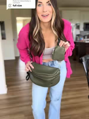 A woman in a bright pink jacket and light jeans stands indoors, holding two bags. The main bag is olive green, the smaller one a matching green. She's smiling at the camera. The setting appears to be a home interior, with a blurred background including a kitchen area. A speech bubble graphic with the words "full size" is in the top left.