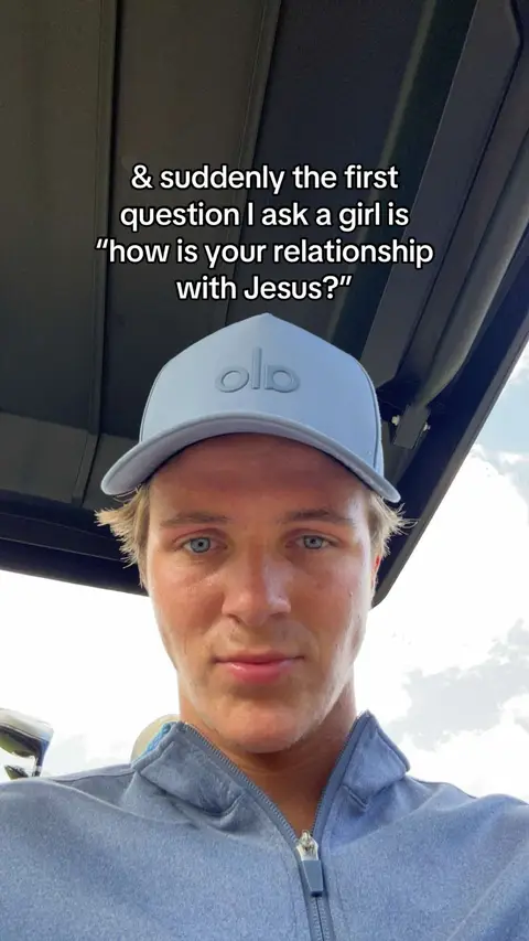 A young man with fair skin and short, light brown hair is centered in the frame, wearing a light blue baseball cap and a matching blue zip-up garment. The background includes a hint of a blue sky and the interior of what seems to be a golf cart, with a dark overhead structure visible. The man is looking directly at the camera with a neutral expression.