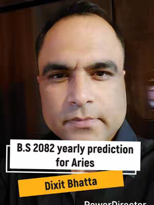 A man with a serious expression is centered in the frame, with text overlays at the bottom. He has short, dark hair and a partially visible, dark-colored button-up shirt. A white rectangle near the bottom holds the text "B.S 2082 yearly prediction for Aries" in dark font, and an orange rectangle beneath it reads "Dixit Bhatta". The background is a blurred indoor setting with a wooden texture.