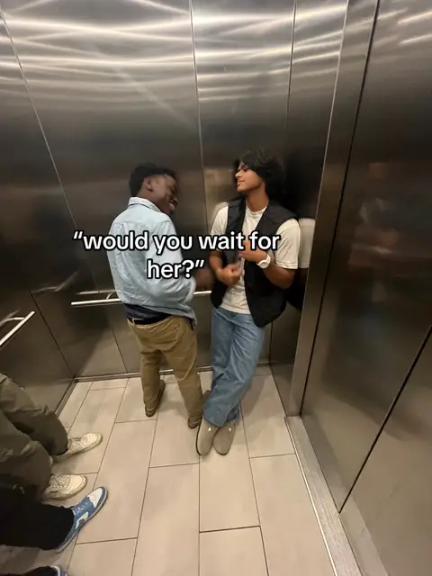 Two young men are in an elevator, one is smiling and the other is looking at him. Text overlay reads, "Would you wait for her?". The setting is a simple elevator with metallic walls and square tiled floor. The two men are casually dressed.