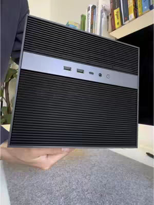 The image shows a black and silver computer device held by a person's hand. The device has horizontal lines across the front with a silver stripe at the top that has several ports and an indicator light. Part of the person's arm and a surface is visible in the background.