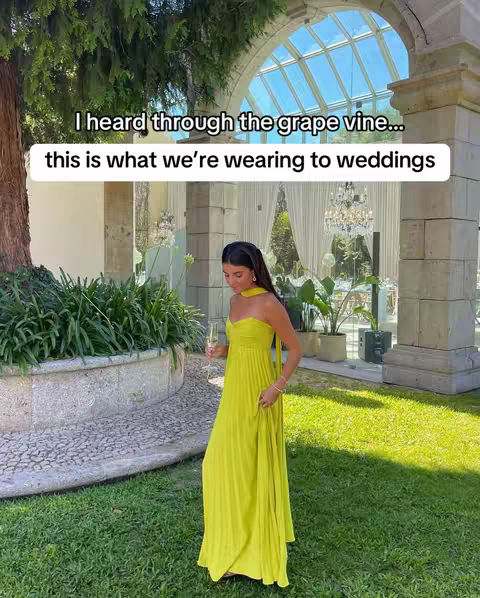 A woman stands in a garden, wearing a bright yellow dress and holding a glass of white wine. A text overlay at the top reads, "I heard through the grape vine... this is what we're wearing to weddings." The scene is set in an elegant outdoor space with formal architecture and greenery.