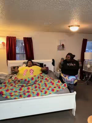 Two women are in a room, where one is lying on a bed with colorful blankets, and the other doing exercise. The room is lit by ceiling lights .