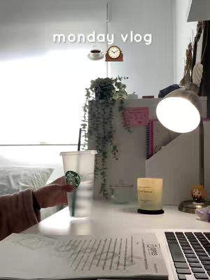 A well-lit desk setup with a laptop, study materials, and a clear Starbucks cup. The top of the image features a text overlay 'monday vlog'.