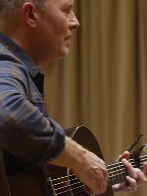 The video shows a man playing an acoustic guitar. He is seen from the side, with his face partially visible, focusing on the instrument and his hands on the fretboard. The background is a simple, light-colored curtain. The man is wearing a blue plaid shirt.