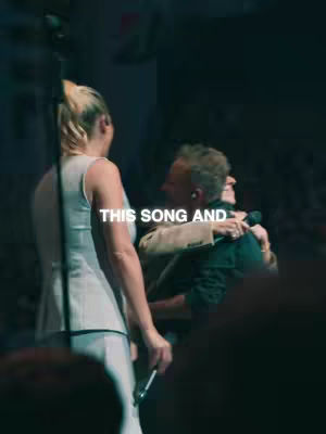 The image shows three people embracing on a stage, captured from a low angle. The foreground features the side view of a woman with blonde hair in a bun, wearing a white dress; she is holding a microphone. A man with a blazer and a man with a dark suit are hugging in the middle ground. The background is dark and blurry, with the lighting focused on the three people. A text overlay reads 'THIS SONG AND'.