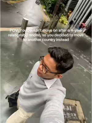 A selfie captured from a high angle, showing a man in the foreground and the exterior environment in the background. The man is wearing glasses and a light-colored sweater, suggesting a cool or casual vibe. The background is a bit blurry, but appears to be an urban setting with a street, some buildings, and a couple of pedestrians. The composition is dynamic, with the foreground elements taking up a large portion of the frame and the background adding context to the scene.
