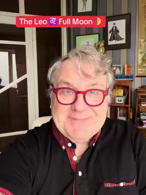 A man with short gray hair and red-framed glasses smiles at the camera, with a red banner that reads 'The Leo Full Moon' across the top. He wears a black shirt with a red patterned collar, and the background includes artwork, a window, and a bookshelf.
