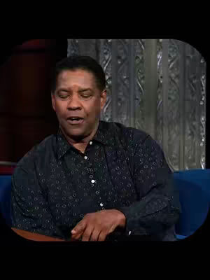 A close-up shot of Denzel Washington, with his mouth open and eyes closed slightly. He is wearing a dark button-up shirt, and the background features patterned, vintage-style decor and a blue couch.