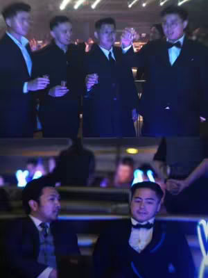 The cover image is a split-screen with two parts. The top portion shows four men, each wearing a black suit and holding a drink, engaging in a toast. The background is blurred with ambient lighting. The bottom portion depicts two other men in similar attire, with one on the left looking at another who wears a bowtie. Their setting is a room with blue lighting. The overall image suggests a gathering or event.