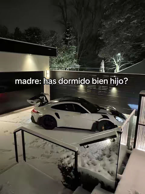 A white sports car is parked outside on a snowy night in front of a modern-style house with a garage and a black fence.