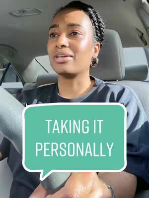 A woman with dark skin is driving a car. She is wearing a black top and gold hoop earrings. Her hair is styled in braids. There is a turquoise speech bubble overlay with the text TAKING IT PERSONALLY in white font. The camera is angled from her head facing forward, with a partial view of the car's interior.