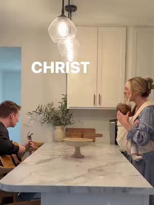 A kitchen scene is captured with a man on the left playing a guitar, and a woman on the right holding a baby. In the center, a marble countertop island dominates the space. Overhead, glass globe lights illuminate the scene. The word 'CHRIST' is superimposed in large, white letters.