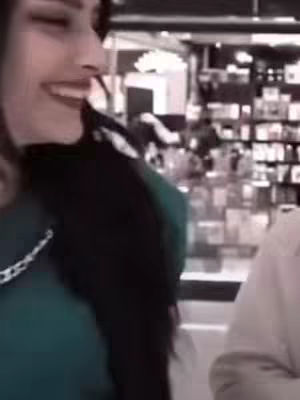 The image shows a woman smiling, looking to her right. She is wearing a dark green shirt. The background is blurry, but appears to be inside a store. The lighting is bright, giving the image a generally high exposure.