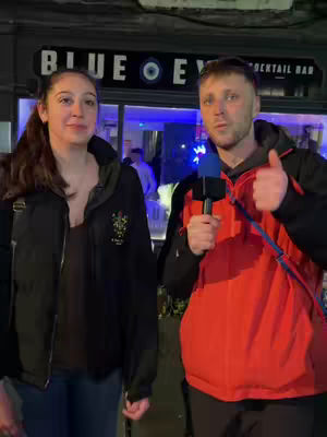 A man holding a microphone and giving a thumbs up while interviewing a young woman in front of a bar entrance. The bar sign says "BLUE EYE COCKTAIL BAR."