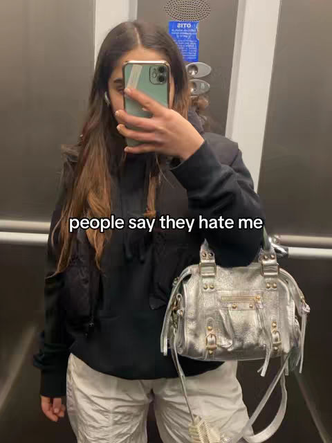 A woman in a black hoodie and white pants stands in an elevator. She is holding her phone up to take a selfie, partially obscuring her face. A silver purse hangs from her arm. The text overlay 'people say they hate me' is at the bottom of the image.