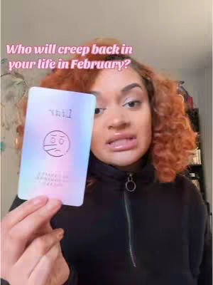 A woman with auburn curly hair holds up a tarot card with a surreal design in front of her. She is wearing a black zip-up hoodie, and the background is out of focus, with a wall and some shelves visible. The text overlay says "Who will creep back in your life in February?"