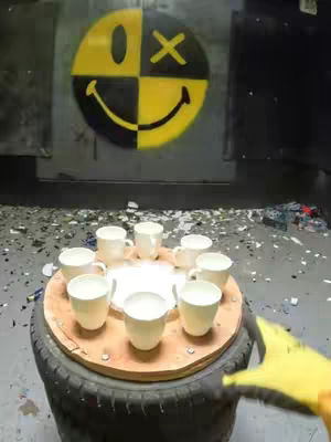 A shot of a table (possibly a tire) with cups on it, in front of a wall that has a yellow and black face design on it. The cup has been placed on it to be smashed by a gloved hand.