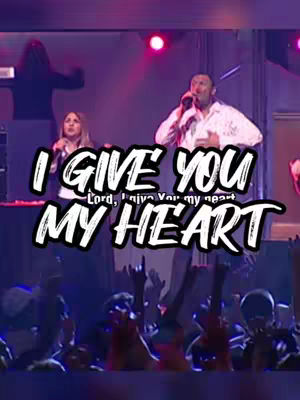 The cover image shows a live performance of a gospel song.  In the foreground, a person sings into a microphone, with a woman at another mic to the  left.  In the background, a silhouette of a person at the drums is visible, and a crowd of people with raised hands is also seen. The text overlay, "I GIVE YOU MY HEART", is prominently displayed in a white block font with a black outline.