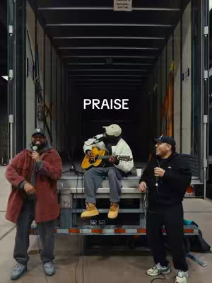 The image shows three men in front of an open cargo container or warehouse bay, with a simple stage or platform set up. The leftmost man wears a red coat and holds a microphone. In the center, another man is seated, playing an acoustic guitar. The third man is on the right, also holding a microphone. The word "PRAISE" is prominently displayed in large white letters across the center of the image. The background is dark and industrial, enhancing the focus on the performers and the message.