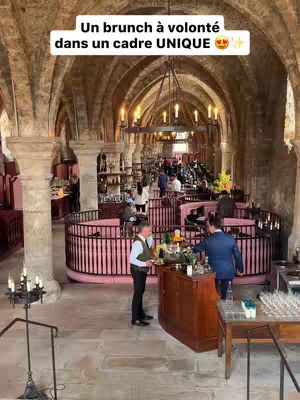 An interior shot of a large, ancient-looking hall. The hall features tall, arched ceilings and stone pillars, with numerous people seated at tables arranged in pink-railed booths. A central bar area has two people standing behind it. Overhead, ornate chandeliers hang from the ceiling, illuminated with warm-toned light. The overall composition creates a sophisticated and welcoming atmosphere.