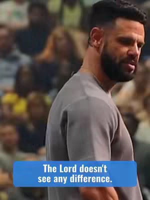 A man with a close-shaved haircut and full beard, dressed in a gray shirt, standing in front of a blurred crowd. A blue box at the bottom contains white text.