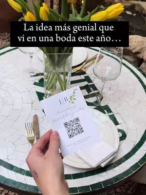 A photo taken from a slightly overhead angle, showcasing a table setting with a floral arrangement. The table, covered in a mosaic pattern, holds a vase filled with yellow tulips. On the table, a menu card with a QR code lies next to a white plate and folded napkin. A hand with a visible tattoo is holding the menu card. In the background, a window and glimpse of outdoor setting are visible, with natural light illuminating the scene.