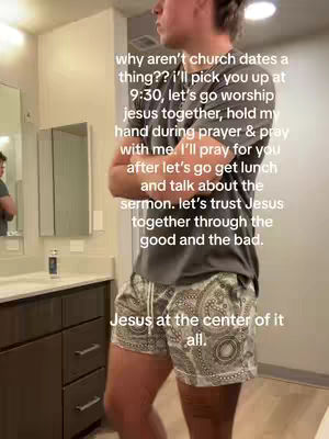 A medium shot of a young man standing in a bathroom, facing slightly right. He has short, styled hair and is wearing a gray t-shirt and patterned shorts. His arms are crossed. The bathroom has neutral-colored walls and a dark wooden vanity with a mirror above it. Text overlays are present, suggesting a message about trust and faith.
