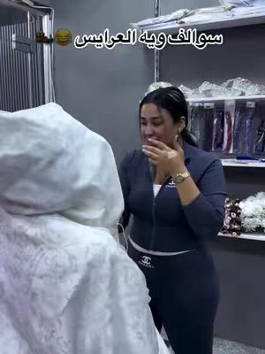 A woman is standing close to a large, white object, possibly a wedding dress. She is looking down with her hand covering her mouth, suggesting a moment of emotion or surprise. She wears a dark blue outfit with a white top, and a silver tiara is on her head, alongside a watch. Behind her are shelves with accessories, possibly in a bridal shop.