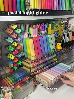 A colorful display of stationery supplies. Organized shelves filled with various highlighters, pens, and markers of different shades (pastels, rainbow, etc.) with some arranged in clear organizers. The composition is bright and visually appealing.